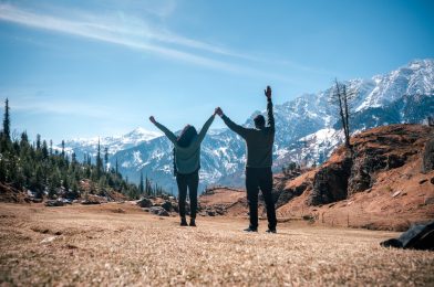 Back view of travelers with raised hands facing a scenic mountainscape, Kothi Meadows, Manali, India